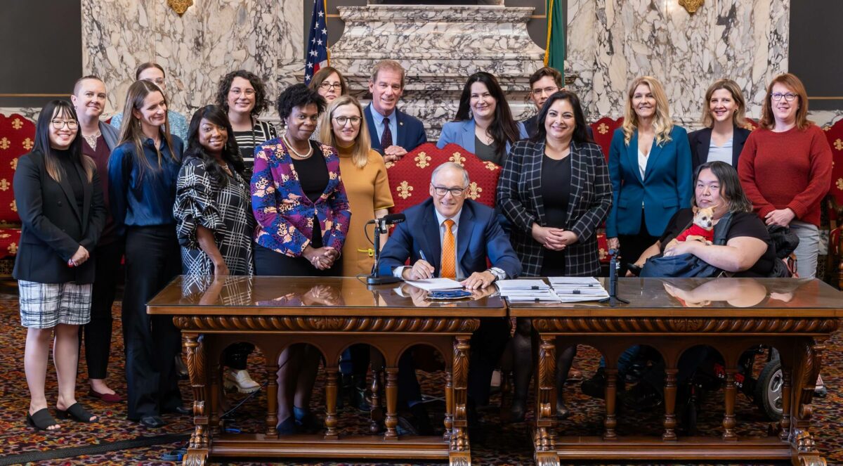 18 Personen stehen hinter dem Gouverneur von Washington, Inslee, der in der Mitte sitzt und einen blauen Anzug und eine orangefarbene Krawatte trägt, und unterzeichnen an einem großen Holzschreibtisch Gesetze.